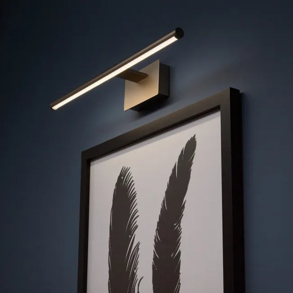 Une lampe murale moderne éclaire avec élégance un tableau encadré sur un mur bleu foncé. La lampe, au design minimaliste, se compose d'une barre lumineuse fine fixée à un support métallique carré, offrant un éclairage subtil et directionnel. Le tableau, aux tons monochromes, présente deux plumes noires sur un fond blanc, encadré d’un cadre noir sobre. L’ensemble crée une ambiance contemporaine et raffinée, idéale pour mettre en valeur une œuvre d’art tout en ajoutant une touche sophistiquée à la décoration 