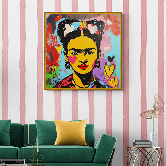 Un tableau street art de Frida Kahlo encadré est suspendu sur un mur à rayures rose et blanc, face à un canapé vert et une table d’appoint dorée, dans un salon au style rétro-chic avec canapé vert et accents dorés.
