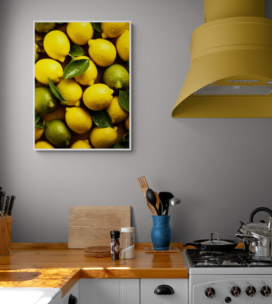 Un tableau de citrons jaunes et verts avec des feuilles vertes est suspendu sur un mur gris dans une cuisine équipée d'un plan de travail en bois et d'un capot de cuisinière jaune vif.